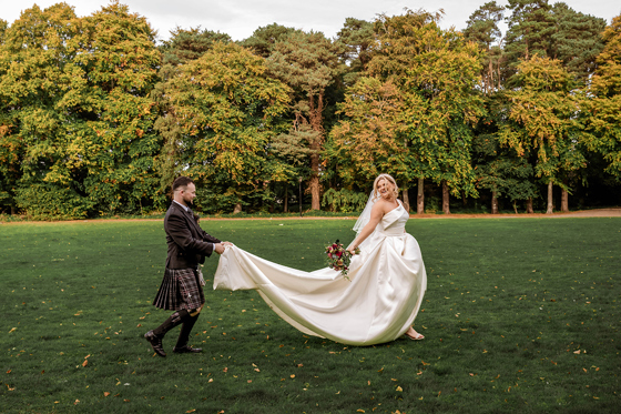 Groom carries his brides train as she runs across grass